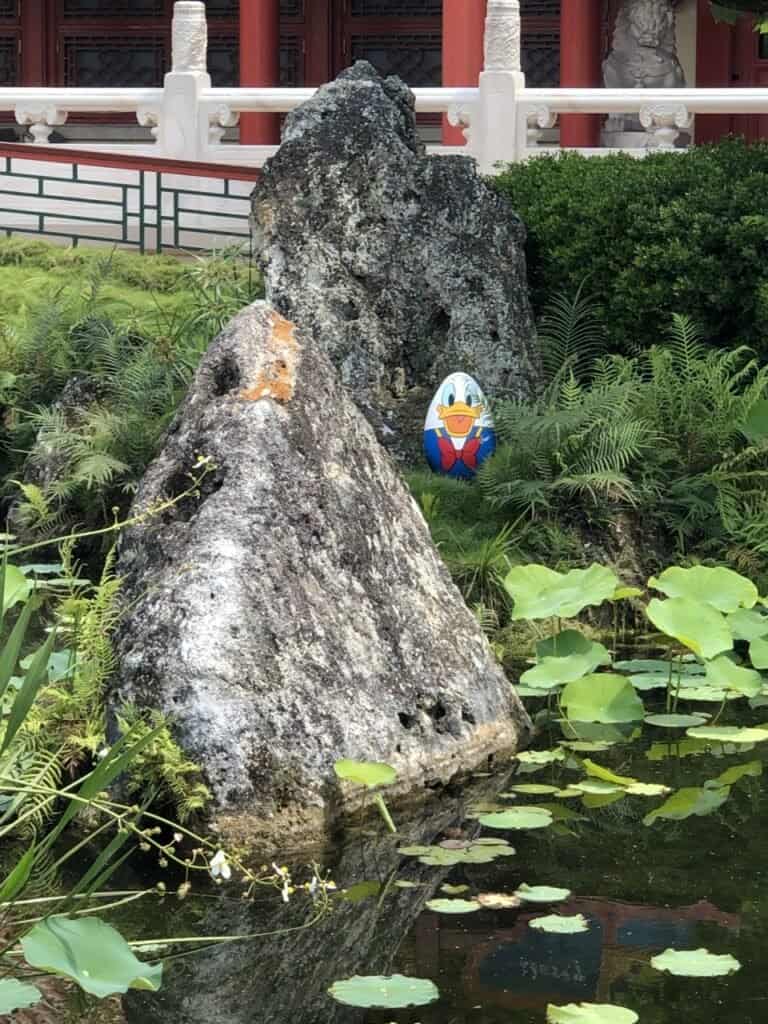 A whimsical Disney Easter egg featuring Donald Duck is nestled among rocks and lush greenery near a tranquil pond with lotus leaves, capturing the festive magic of springtime at Disney Parks.