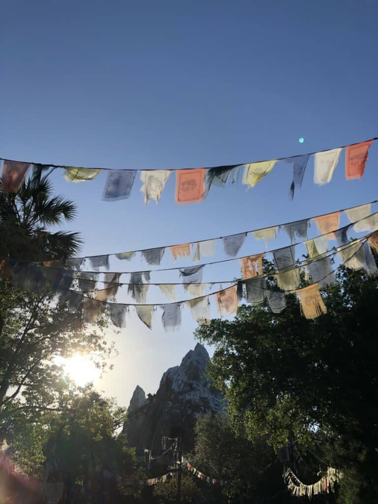 Vibrant prayer flags flutter across a brilliant blue sky above the lush landscapes, sunlit mountains, and towering trees of Disney’s Animal Kingdom, perfectly capturing the park’s lively atmosphere and adventurous spirit.