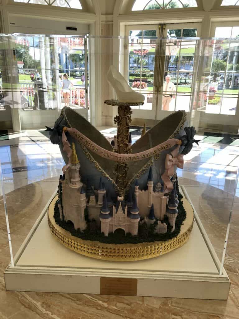 A magical Disney World-inspired cake designed as Cinderella Castle and her iconic glass slipper, showcased in a glass display case under bright lights.