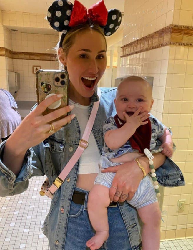 A Disney fan sporting Minnie Mouse ears snaps a joyful mirror selfie in a tiled restroom, grinning alongside her happy baby after an epic day at Magic Kingdom—capturing the magic just moments before park closing.