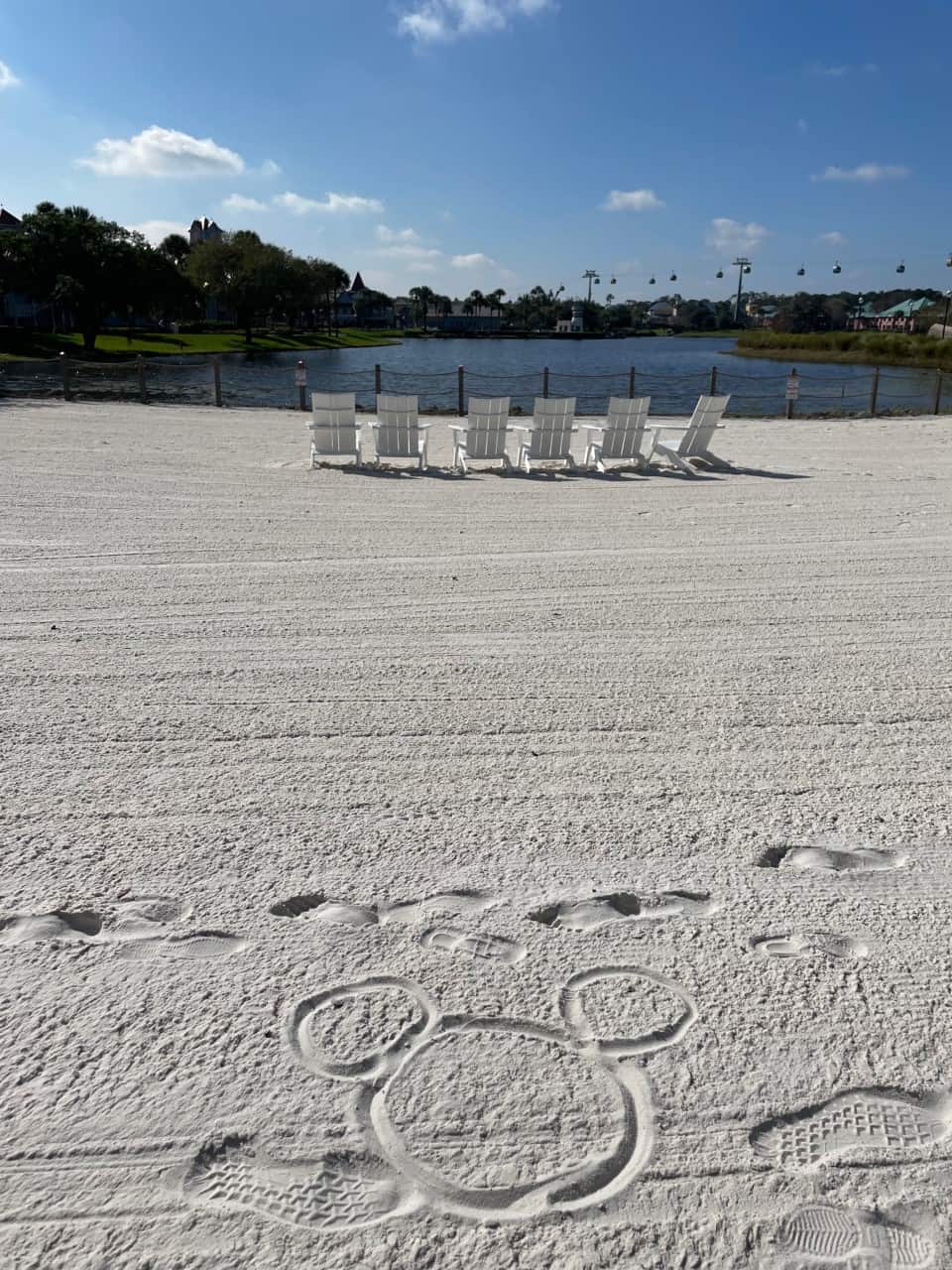 Sun-soaked white sand beach at Riviera Resort, lined with inviting chairs beside sparkling blue water and under a clear sky. A whimsical Mickey Mouse head is artfully drawn in the sand—a signature Disney touch that adds extra magic to this resort experience.