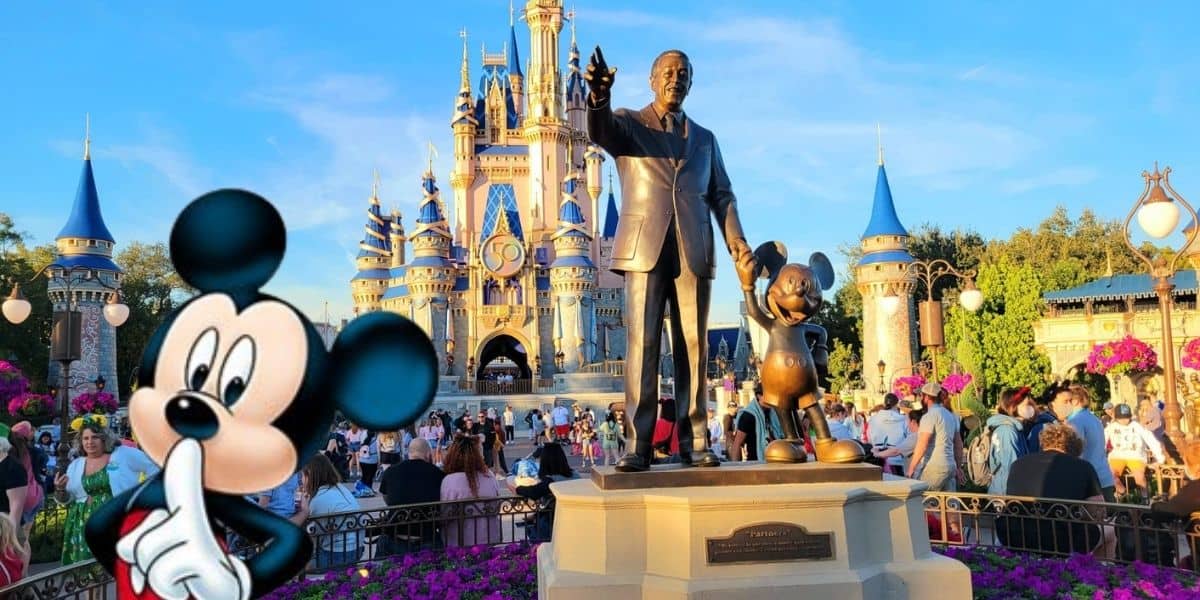 A statue of a man holding hands with a mouse character stands prominently in front of a fairy-tale castle. Amidst vibrant flowers and lush greenery, visitors enjoy the sunny day while considering Disney money-saving tips like arriving early for the best parking spots.