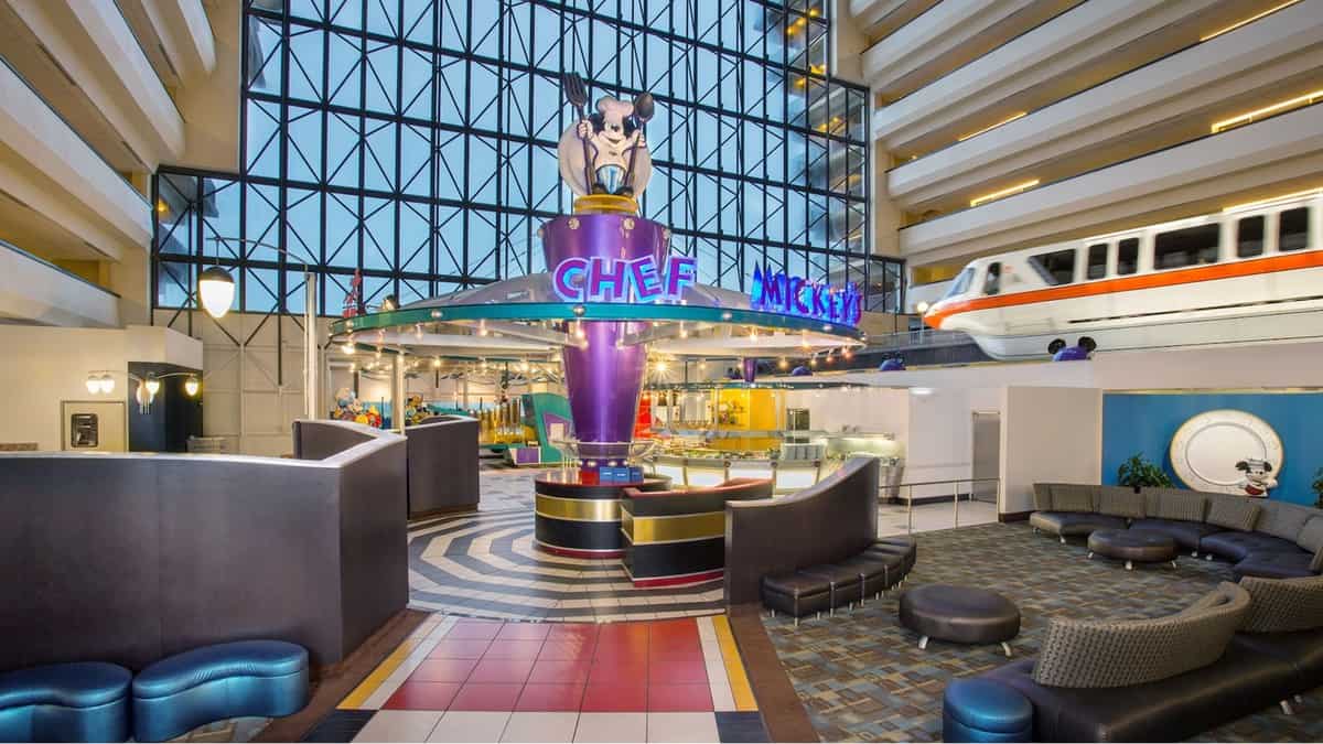 Step into the vibrant world of Chef Mickey’s at Walt Disney World, where a smiling Mickey Mouse statue enthusiastically waves to guests amid playful curved seating, all set against the iconic backdrop of a monorail gliding by.