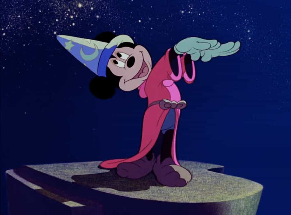 Mickey Mouse dressed as the Sorcerer's Apprentice stands on a rock with his arm raised, surrounded by the enchanting starry night sky of Fantasia Gardens at Disney World.
