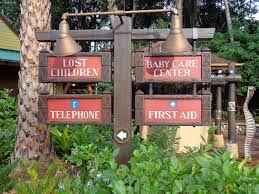 Charming wooden signpost at Disneyland points guests to Lost Children, Baby Care Center, public telephones, First Aid station, and Disney Doctor services amid vibrant landscaping.