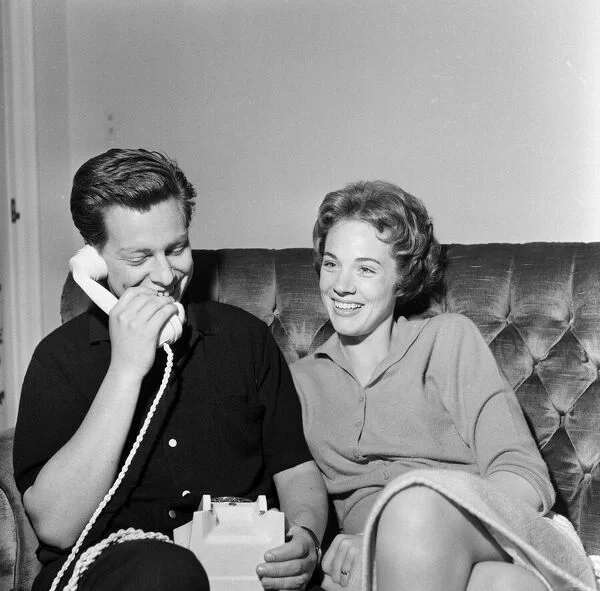 A smiling man chats on the phone while sitting with a woman on a couch; both appear cheerful and at ease, as if reminiscing about magical moments from Broadway productions or discussing legendary set designer Tony Walton’s passing at 87—a black and white image that captures their shared love of theatre and classic entertainment.