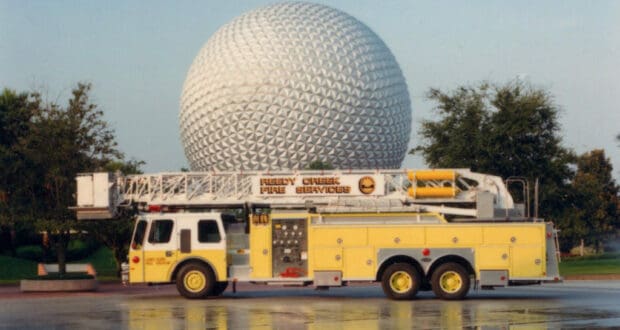 A bright yellow Reedy Creek fire truck is parked right in front of EPCOT’s iconic Spaceship Earth at Walt Disney World, with glistening wet pavement and lush trees in the background.