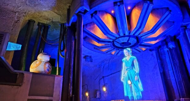 A lifelike hologram of Rey stands next to BB-8 in a high-tech Star Wars setting filled with vibrant blue and orange lights, transporting guests straight into the action of Disney World’s Rise of the Resistance ride—where, in this scene, every effect works flawlessly for an uninterrupted adventure.