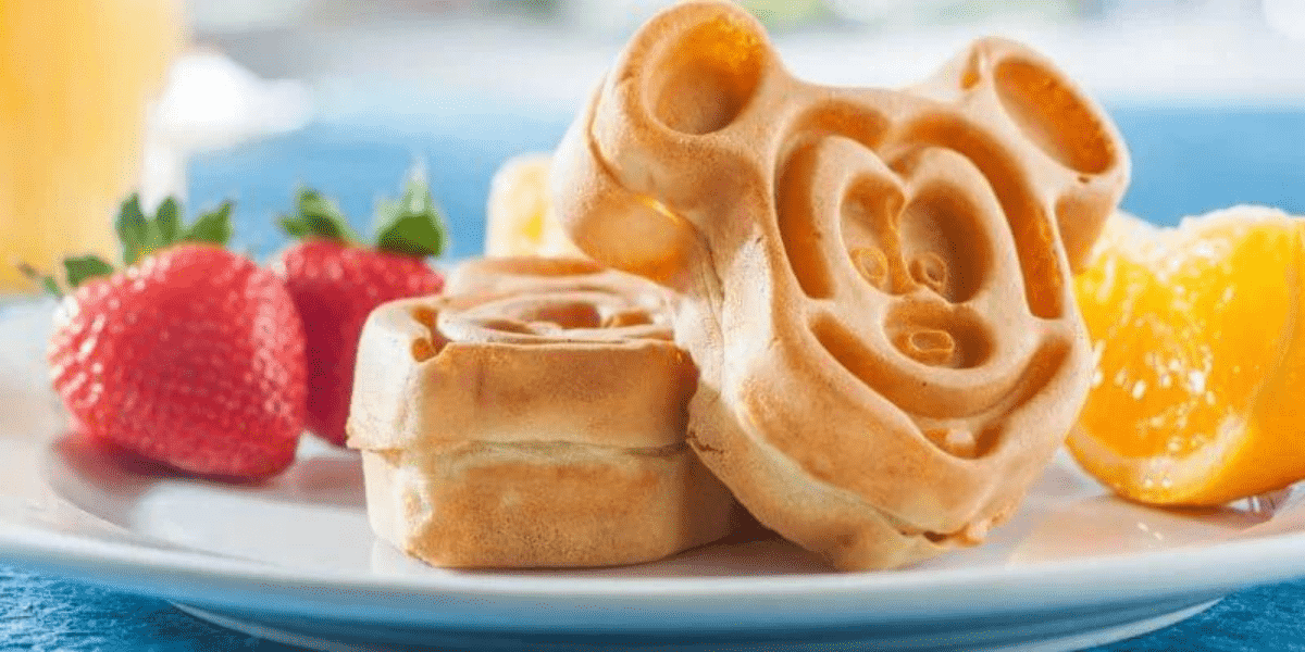 A plate with waffles shaped like a cartoon character, accompanied by fresh strawberries and orange slices, sits atop a blue tablecloth—a delightful Disney Dining experience perfect for breakfast.