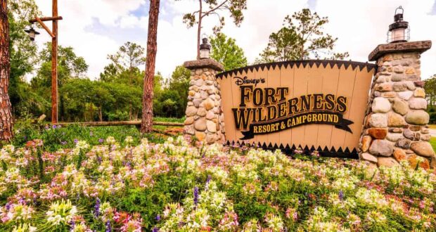 A large wooden and stone sign reading "Disney's Fort Wilderness Resort & Campground" stands behind a bed of colorful wildflowers, inviting guests to explore Disney Parks and discover hidden secrets among tall trees and lush greenery.