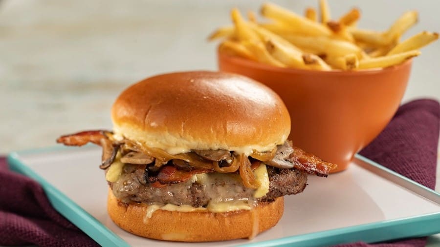 Savor a mouthwatering burger loaded with cheese, mushrooms, onions, and bacon on a soft bun, paired with golden fries in the background—perfect for refueling during your day of adventure at EPCOT’s Connections Eatery. Enjoy flavorful bites in the heart of park excitement at family-friendly prices.