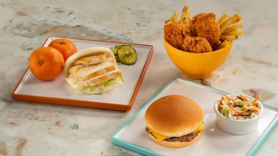 Three plates of food showcase EPCOT Connections Eatery pricing: sliced grilled chicken on a bun with tangerines, fried chicken tenders with French fries, and a cheeseburger paired with a small serving of coleslaw.