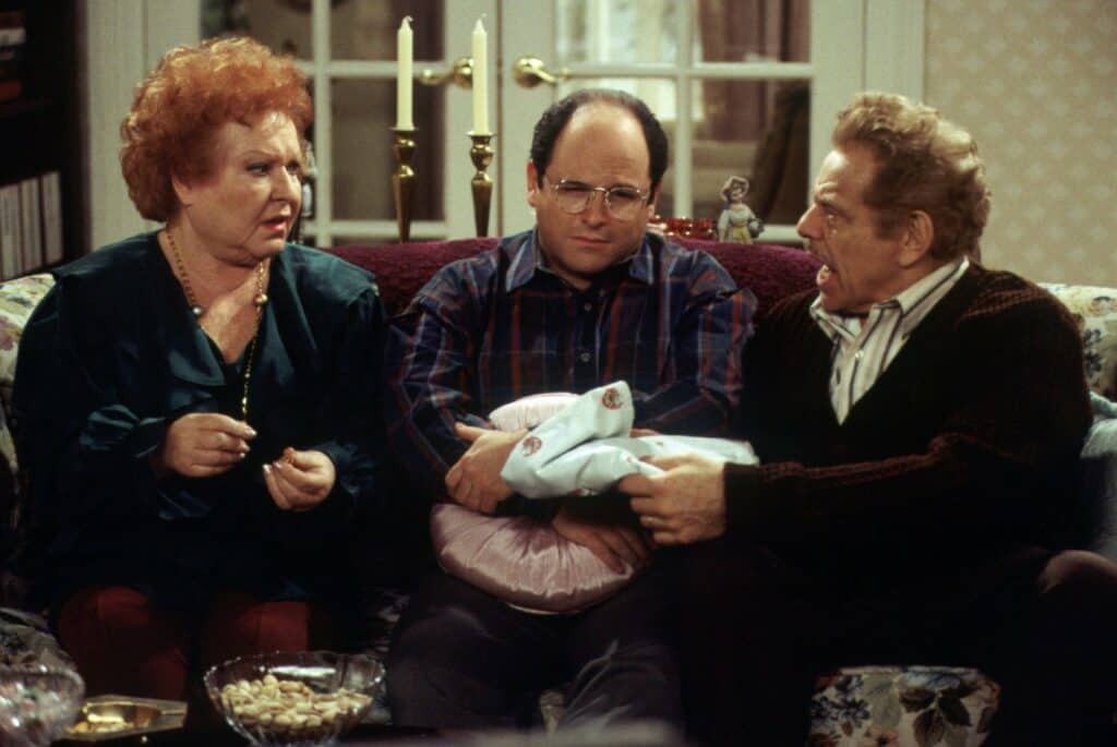 Three fans relax on a lounge inside a themed entertainment venue; the guest in the center clutches a souvenir package and appears uneasy, possibly reacting to an announcement about Estelle Harris’s passing displayed on screens nearby.