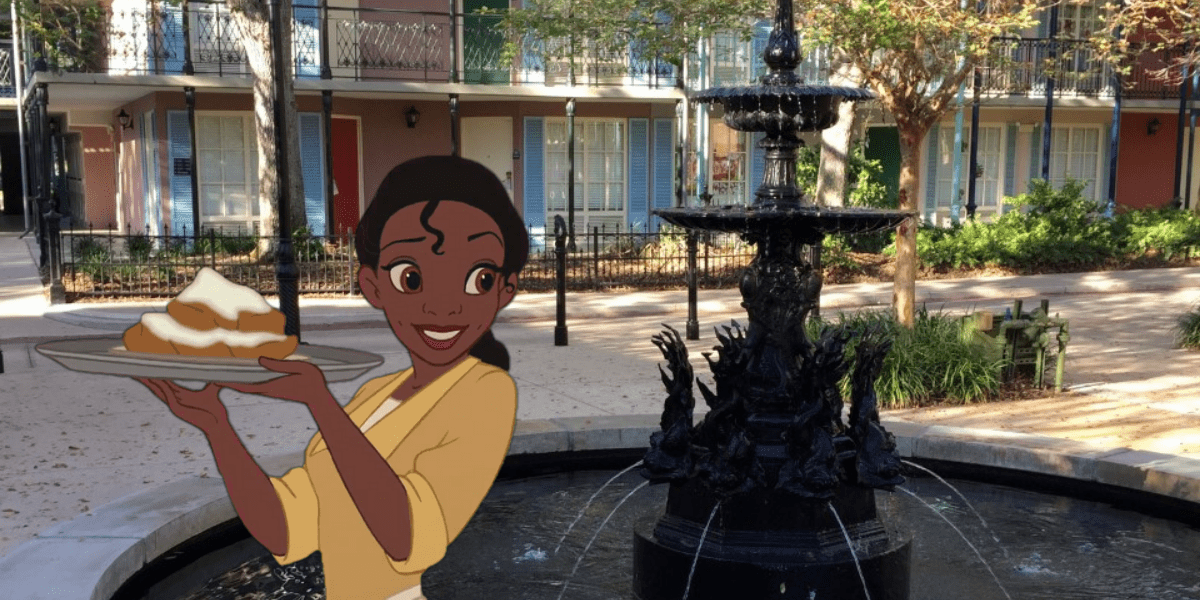 Tiana enjoying a powdered-sugar beignet beside the black fountain in the vibrant, sunlit courtyard of Disney’s Port Orleans Resort, surrounded by lush trees and classic New Orleans-inspired buildings.