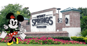 Minnie Mouse shows off her latest Disney Springs shopping spree, holding colorful bags full of Disney merchandise in front of the iconic welcome sign and surrounded by bright, cheerful flowers.