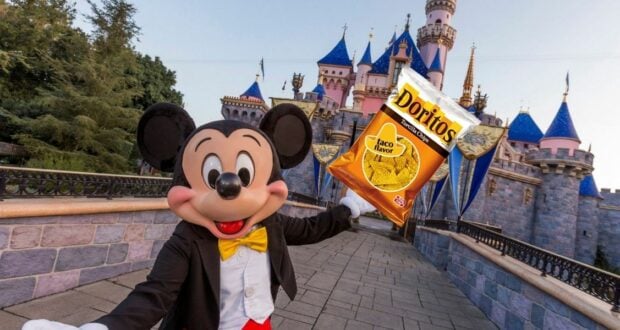 Mickey Mouse stands in front of Sleeping Beauty Castle at Disneyland, holding a bag of Doritos and captivating an enthusiastic crowd with fun Disney World trivia.