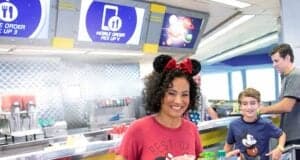 A smiling woman wearing Mickey Mouse ears and a “Best Day Ever” shirt carries food trays in a modern restaurant, with a boy beside her and Disney World Mobile Ordering pick-up signs glowing in the background.