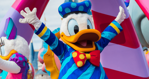 Donald Duck, dressed in his classic blue sailor suit and red bow tie, energizes the crowd with arms raised during a vibrant Disney Parades celebration at Disney Resorts near Fort Wilderness, while elaborately decorated golf carts roll by in the background.