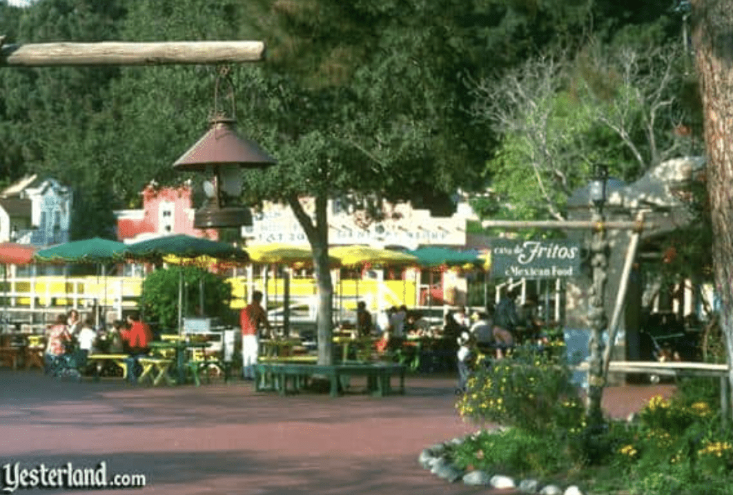 Vibrant patio tables shaded by colorful umbrellas invite guests to relax and dine beneath lush trees, with the classic Casa de Fritos Mexican food sign nearby—a favorite Disney World spot to unwind and soak up park history.