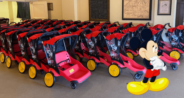 Walt Disney World strollers in classic red and black designs featuring Mickey Mouse line up, with a cheerful cartoon Mickey Mouse welcoming guests at the front.