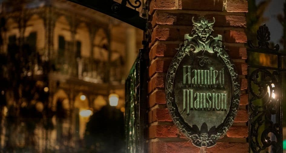 The iconic bronze Haunted Mansion sign, featuring a nod to the legendary Hanging Scene, stands atop a brick pillar as guests approach the eerily illuminated facade of the Haunted Mansion at night.