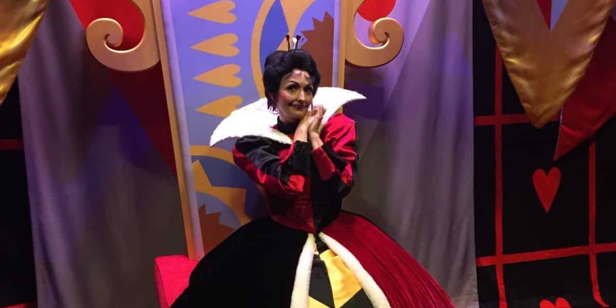 In front of a vibrant backdrop adorned with heart motifs, a person dressed as the Queen of Hearts from "Alice in Wonderland" channels the spirit of Disney Villains. Their red and black costume, complete with a white collar and elaborate headpiece, would fit right into Club Villain at Disney's Hollywood Studios.