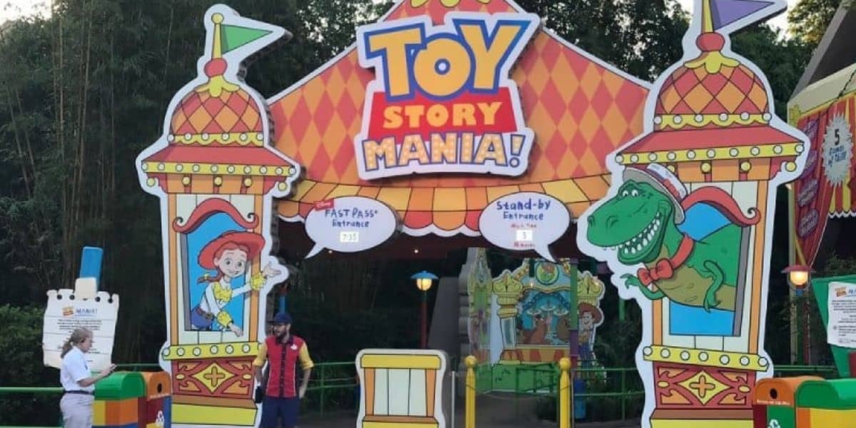 The entrance to Toy Story Midway Mania features colorful decorations with characters Jessie and Rex. Two staff members stand near the entrance under a large sign with flags and playful designs, inviting guests to the FastPass+ and Stand-by lines, ready to reveal the secret to achieving a high score.