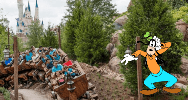 Guests experience the excitement of a Disney roller coaster near Cinderella Castle, while Goofy poses playfully among the trees in the foreground, appearing to assist fans in a whimsical search for a lost item.