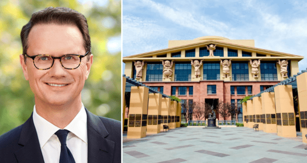 An executive in a sharp suit and glasses stands beside an image showcasing a Disney attraction, featuring grand statues on its upper facade—an architectural nod to the imaginative landmarks Peter Rice helped bring to life at Disney parks.