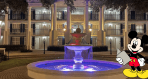 Mickey Mouse stands in the foreground holding a map, welcoming guests outside a Disney Resort hotel at night, with a beautifully lit fountain enhancing the magical atmosphere.