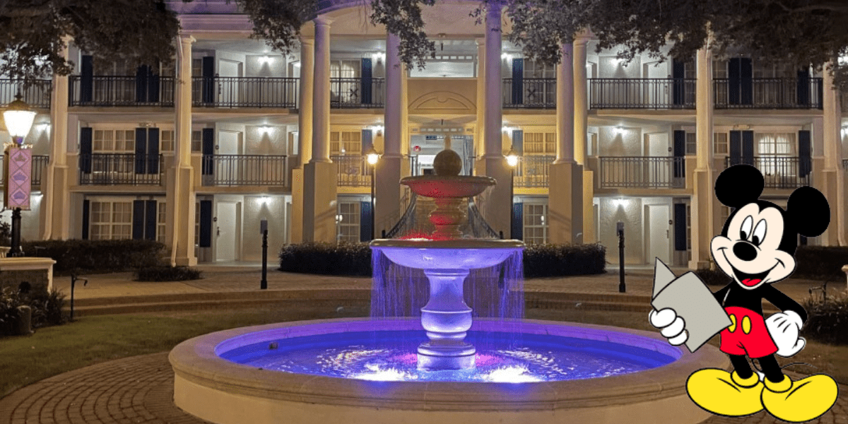 Mickey Mouse stands in the foreground holding a map, welcoming guests outside a Disney Resort hotel at night, with a beautifully lit fountain enhancing the magical atmosphere.