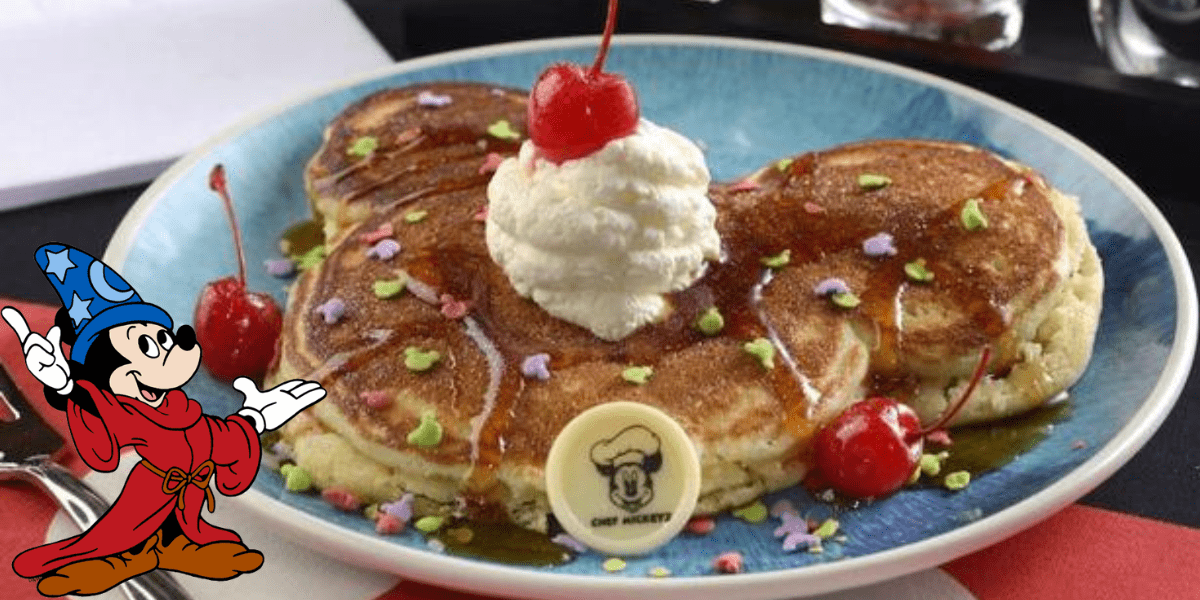 A plate features three pancakes arranged in the shape of a famous mouse's head, topped with whipped cream and a cherry. Colorful sprinkles decorate the pancakes, alongside cherries and a white chocolate piece with a chef hat design. It's the perfect Mickey snack for any Disney Dining experience, complete with a wizard character on the left.