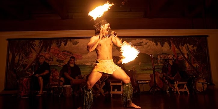 A fire dancer on stage at the Universal Wantilan Luau Dinner Show, twirling flaming batons with live musicians seated behind, capturing the excitement and energy of theme park evening entertainment, much like the legendary performances at Disney’s Spirit of Aloha.