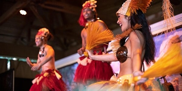 Performers in vivid costumes showcase a lively traditional Polynesian dance onstage, capturing the festive spirit of both the Universal Wantilan Luau Dinner Show and Disney's Spirit of Aloha at their respective theme park resorts.