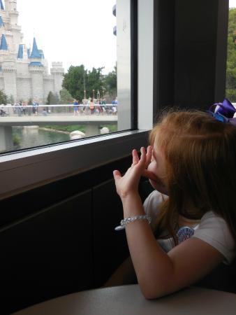 A young girl sporting a signature Minnie Mouse bow gazes out the window toward Cinderella Castle, waving enthusiastically as she dreams of all the magical adventures—and maybe even her favorite Disney Dining Tips—awaiting her on her royal visit.
