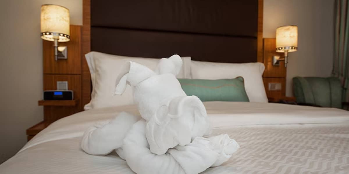 A towel folded into the shape of a dog sits on a neatly made bed with white linens in a Disney Cruise Verandah Stateroom, two pillows, a green cushion, and two lit bedside lamps completing the cozy scene.