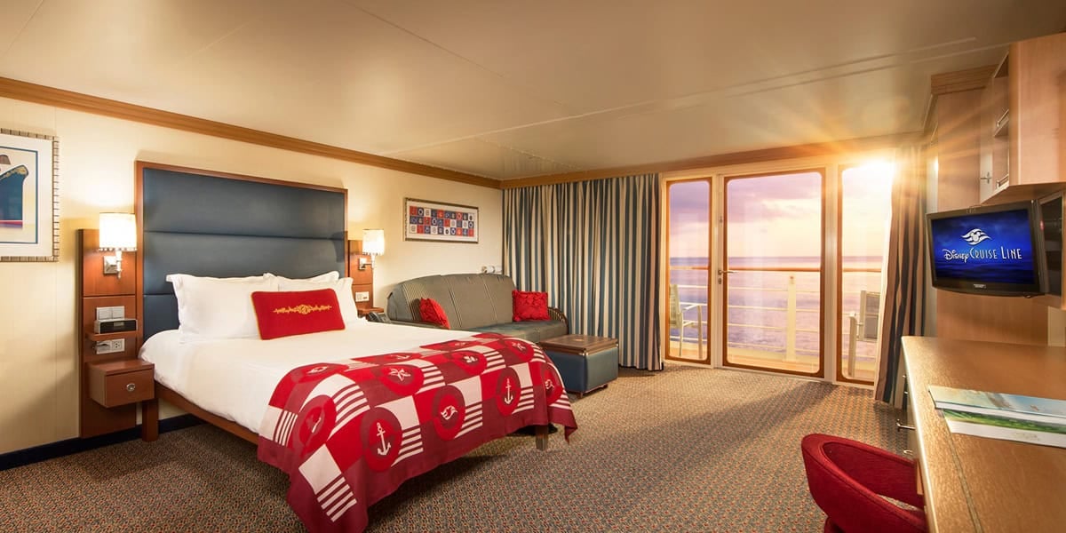 A bright, spacious hotel room styled like a Disney Cruise Verandah Stateroom, featuring a large bed with a red nautical-themed blanket, gray sofa, desk with TV, and glass doors opening to a balcony with an ocean view at sunset.