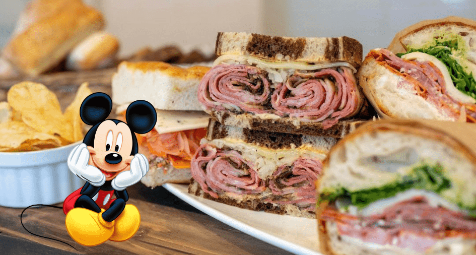 Mickey Mouse at Disney World sits beside a tempting plate of assorted sandwiches and chips from BoardWalk Deli, all set on a wooden table.