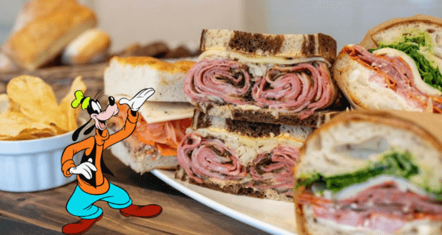 Goofy posing by a tempting spread of piled-high deli sandwiches and crispy potato chips, radiating the lively, whimsical atmosphere of Disney's BoardWalk Deli.