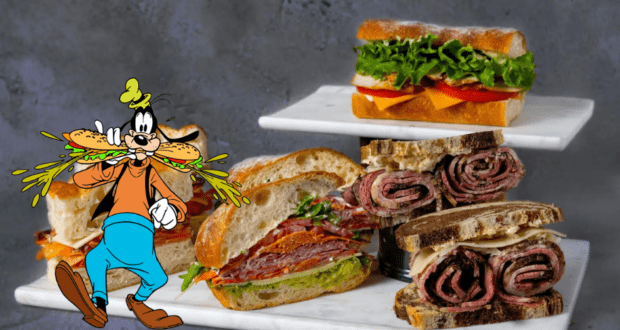 Goofy shows off a giant sandwich beside a tempting tiered display of deli creations on marble stands inside the lively BoardWalk Deli at Walt Disney World.