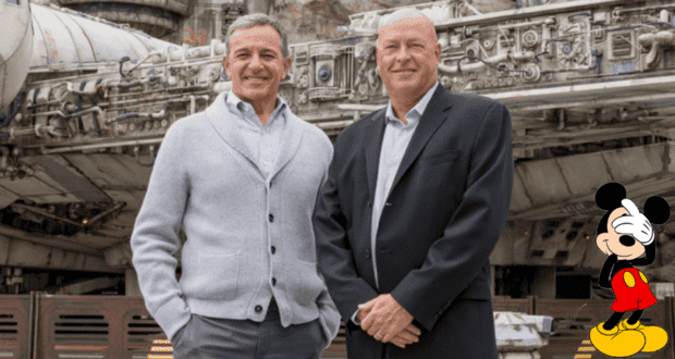 Disney executives Bob Iger and Bob Chapek, dressed in suits, smile for a photo in front of a space-themed attraction, while Mickey Mouse appears in cartoon form at the bottom right corner.
