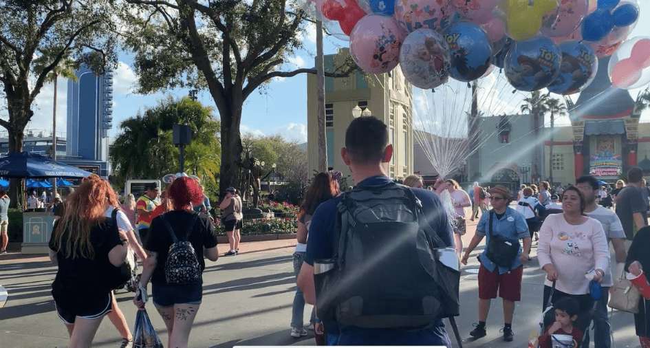 Guests stroll through Disney World, sporting backpacks filled with must-have essentials, as colorful Mickey balloons float above under the bright Florida sun.