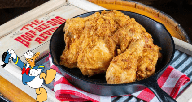 Donald Duck welcomes guests beside the iconic "Hoop-Dee-Doo Revue" sign, with a skillet of golden fried chicken resting on a red-checkered cloth, capturing the lively flavors and festive spirit of Disney’s Hoop-Dee-Doo Musical Revue.