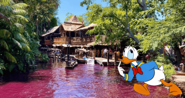 Donald Duck by the Jungle Cruise’s pink-hued river, posing near a rustic wooden cabin nestled among lush green trees in Adventureland.
