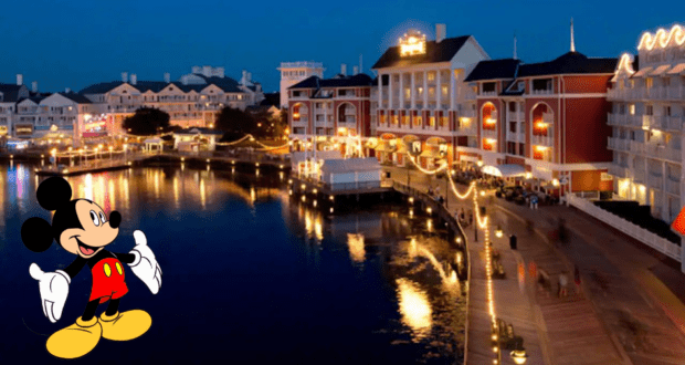 Mickey Mouse greets guests along the glowing lakeside at Disney's BoardWalk during dusk, with the vibrant lights of the restaurant and boardwalk reflecting across the shimmering water.