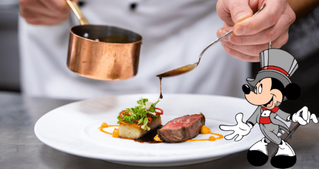 At Victoria & Albert's in Disney World, a chef skillfully plates a gourmet steak dish with sauce as Mickey Mouse, dressed smartly in a suit, stands beside the plate overseeing the culinary magic.