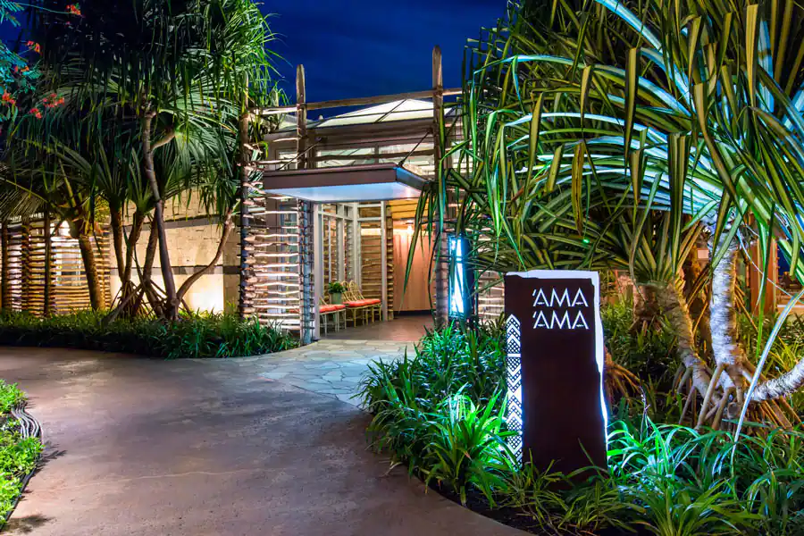 Nighttime view of the AMA AMA restaurant entrance at Disney's Aulani Resort, with glowing 'AMA AMA' signage and vibrant tropical landscaping welcoming guests.