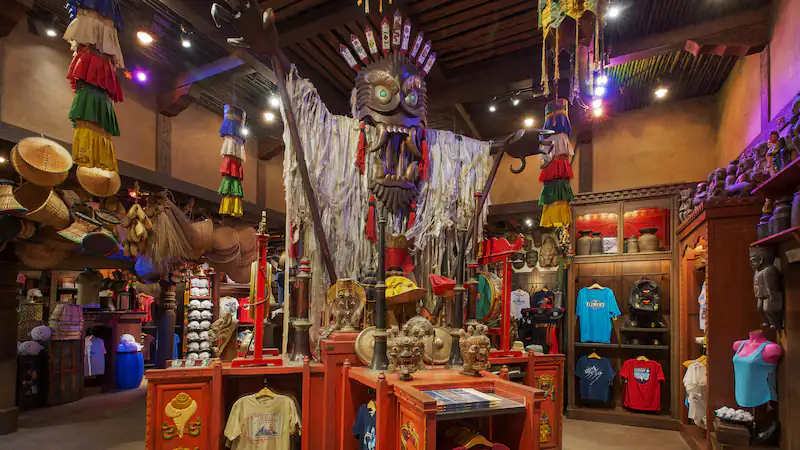 A vibrant souvenir shop in Animal Kingdom style, bursting with tribal-inspired decor, rows of colorful Disney tees and hats, all centered around a towering tiki statue perfect for iconic theme park photo ops.