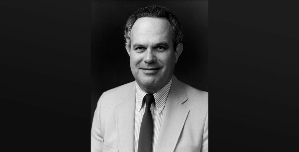 Black-and-white portrait of Disney Legend Ron Logan, the visionary behind countless Disney Parks entertainment spectaculars, smiling in a light suit, striped shirt, and tie.
