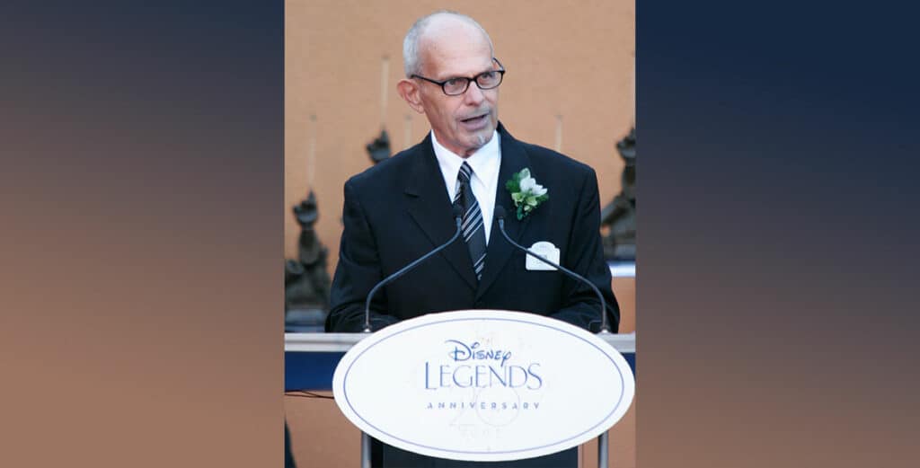 Ron Logan, Disney Legend and creative force behind beloved park entertainment, stands at the Disney Legends podium in a sharp suit and signature glasses, accented by a flower on his lapel, addressing fellow fans and cast members during the iconic ceremony.